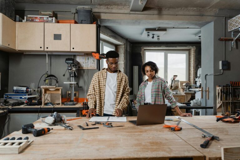 Two people collaborating on a woodworking project in a modern workshop, using tools and a laptop.